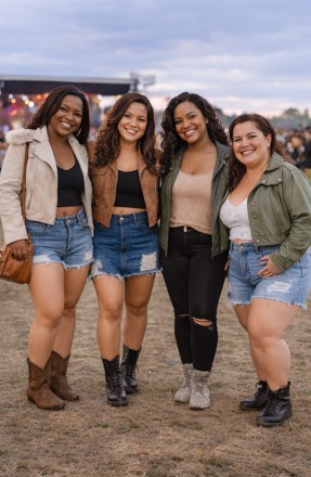 Four friends in denim shorts and boots happily pose for a picture outdoors.