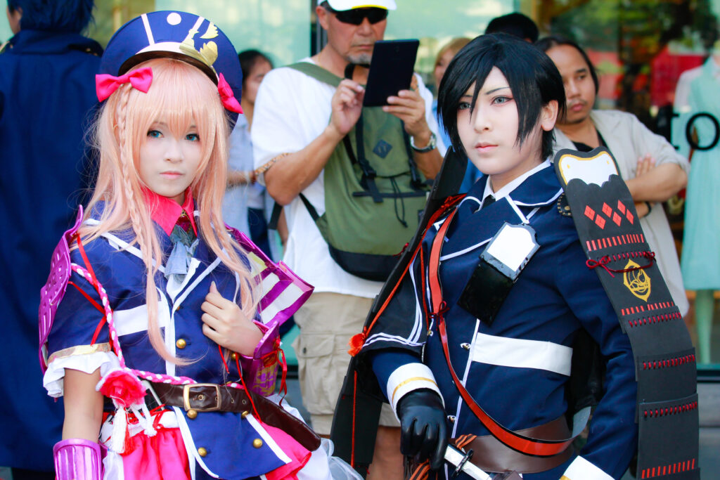 Two cosplayers in detailed anime military style costumes posing at an anime convention with colorful wigs and elaborate accessories.