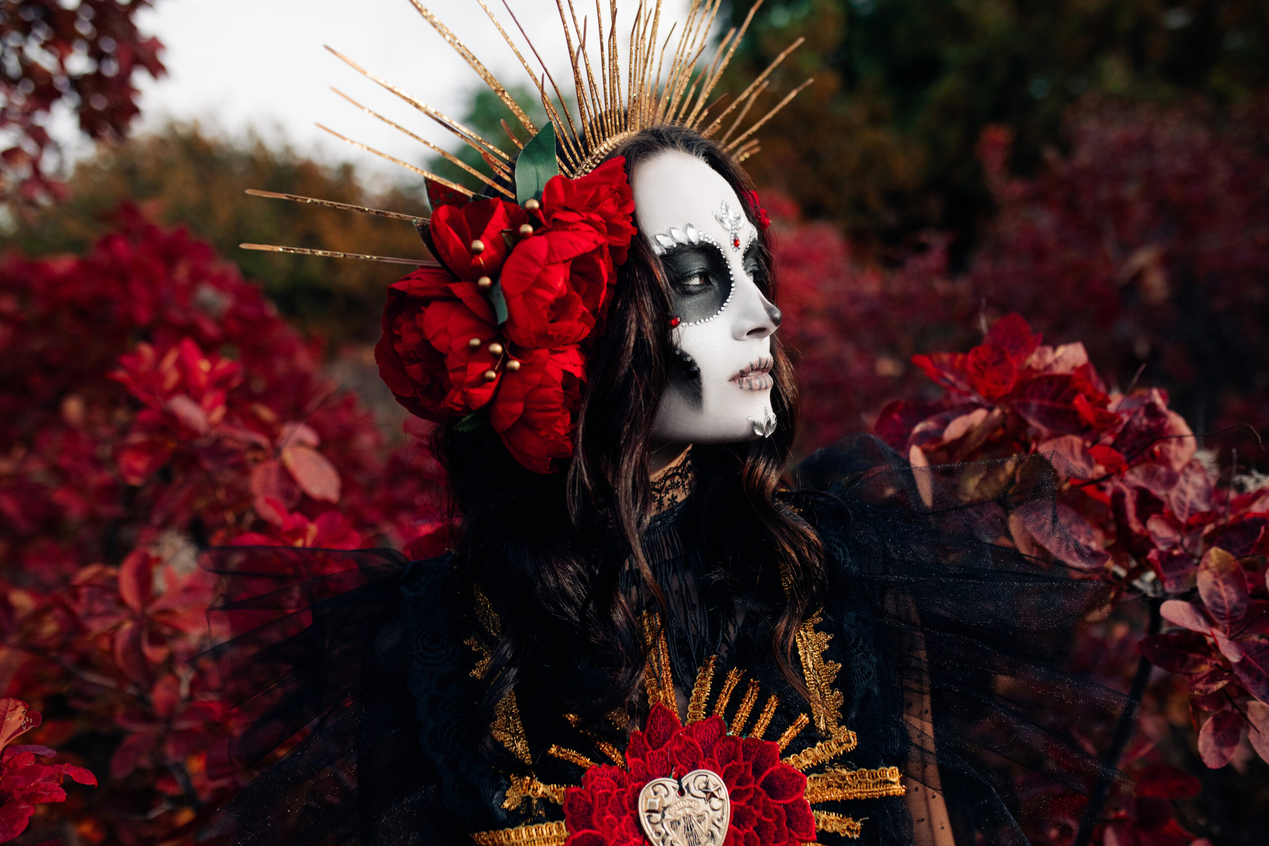 Woman in gothic costume with dramatic skull face paint, red flowers, and golden crown headpiece in a dark autumn forest setting.