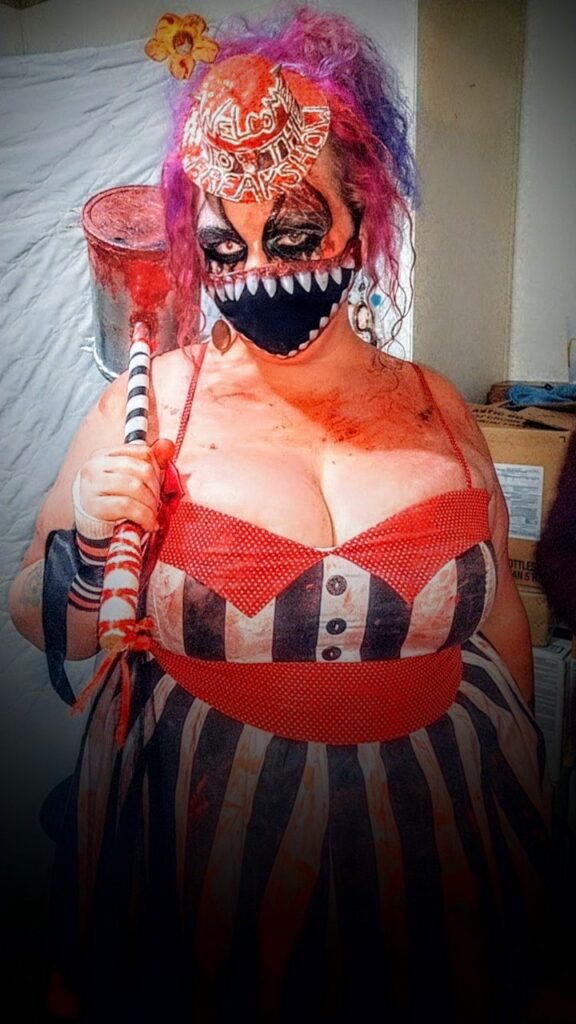 Plus size woman in a creepy freakshow clown costume with striped dress, colorful hair, and horror mask holding a carnival hammer at a haunted house attraction.
