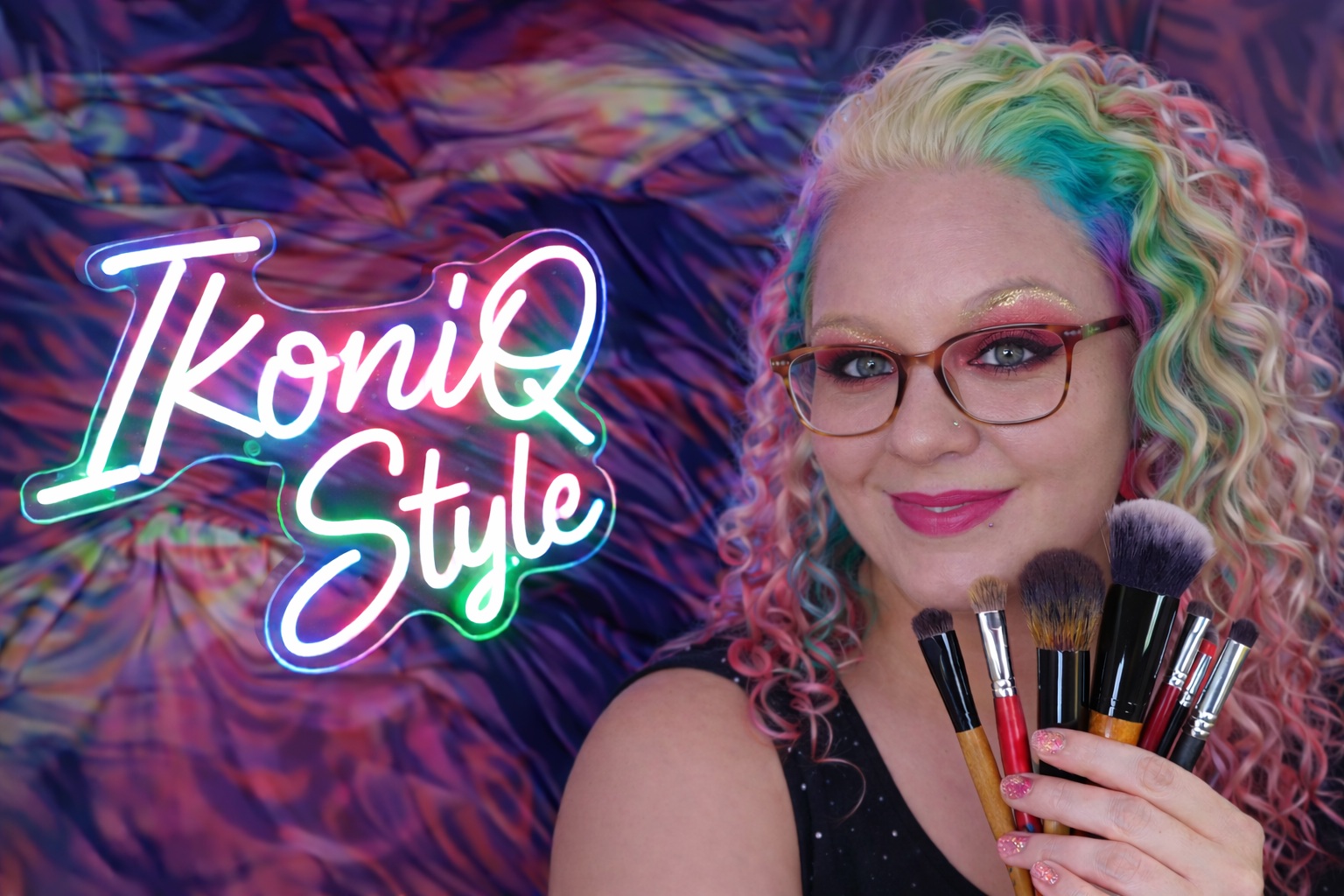 Woman with rainbow-toned curly hair and glasses smiling while holding a variety of makeup brushes, posing in front of a colorful neon “IkoniQ Style” sign and vibrant abstract background.