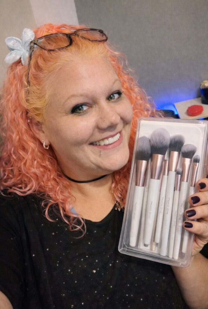 Woman with pink curly hair holding a full set of makeup brushes from EIGSHOW in clear packaging, smiling while showcasing the product.