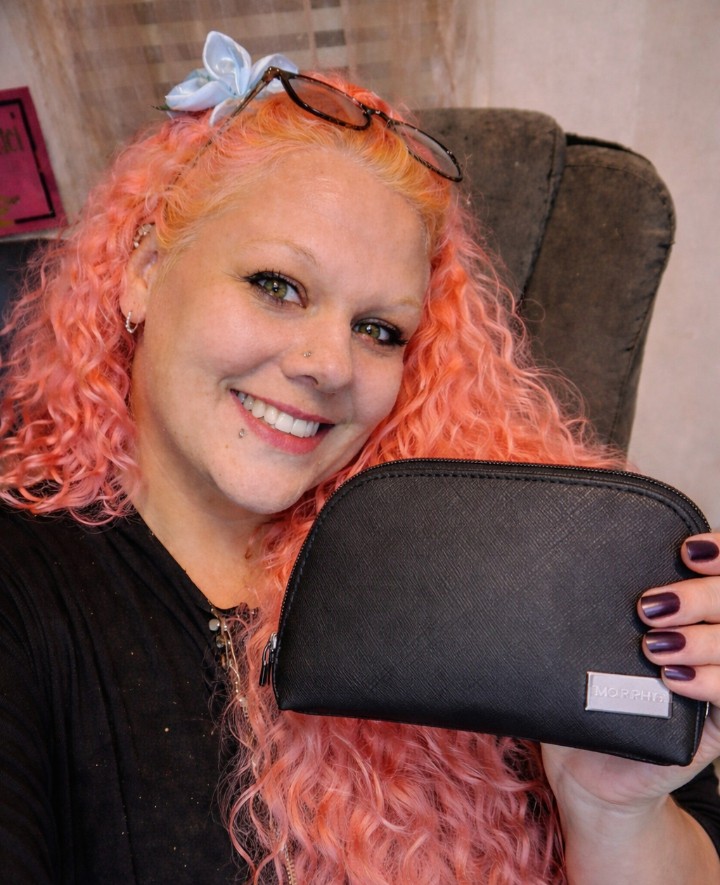 Woman with pink curly hair holding a black Morphe makeup bag, smiling and posing in a cozy indoor space.