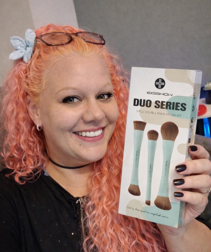 Woman with pink curly hair holding an EIGSHOW Duo Series makeup brush set box, smiling at the camera in a relaxed home environment.