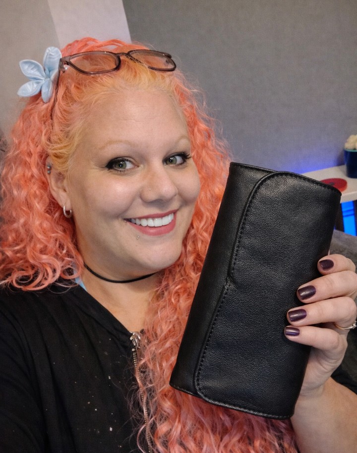 Woman with pink curly hair smiling while holding a black makeup brush case, wearing glasses on her head and a blue flower hair clip in a casual indoor setting.