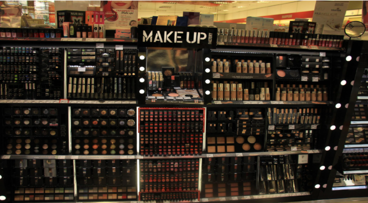 A well-organized makeup display inside a retail store, featuring shelves filled with foundations, lipsticks, eyeshadows, and powders. A central section labeled “MAKE UP” is surrounded by illuminated mirrors, with rows of products arranged by type and shade. The display includes testers and boxed items, with a wide variety of tones and colors neatly lined across multiple tiers.
