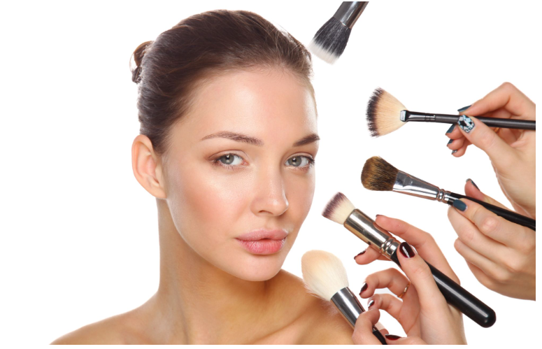 A close-up portrait of a woman with natural, glowing makeup and her hair pulled back, while multiple hands hold different makeup brushes near her face, demonstrating application techniques against a clean white background.