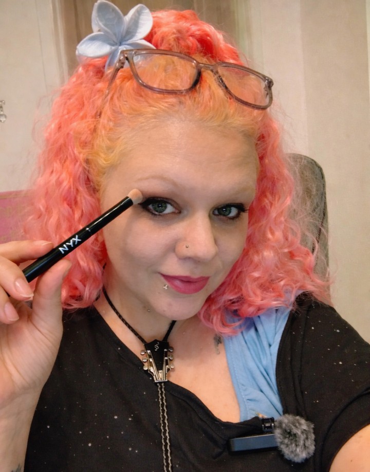 Woman with pink curly hair demonstrating a NYX precision eye brush near her eye, showing how to apply detailed eyeshadow.