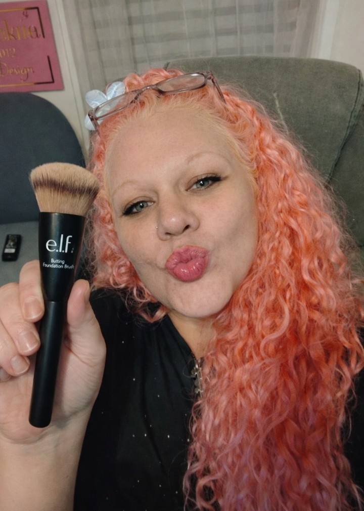 Woman with pink curly hair making a playful kissy face while holding an e.l.f. Buffing Foundation Brush close to the camera in a casual indoor setting.
