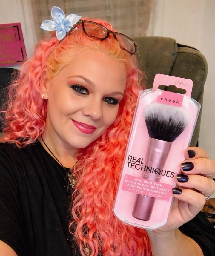 Woman with pink curly hair holding a Real Techniques mini multitask brush in pink packaging, smiling and presenting the product to the camera.