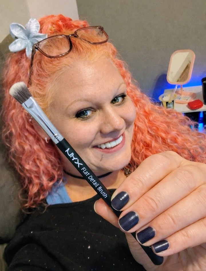 Woman with pink curly hair smiling while holding a NYX Flat Detail Brush close to the camera, highlighting the brush shape and detailing.