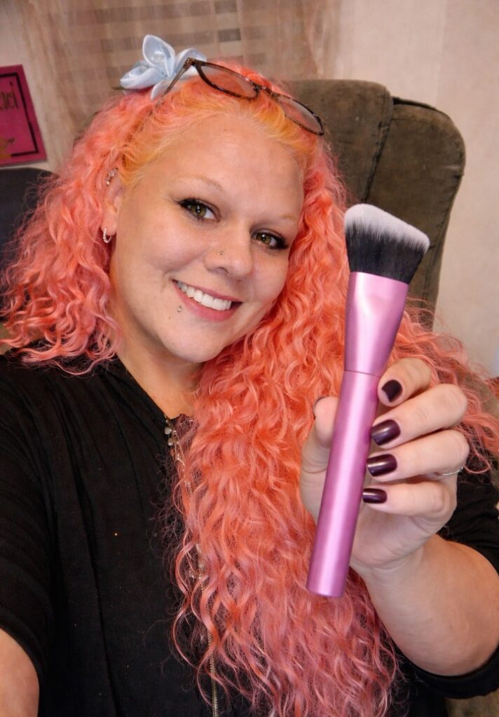 Woman with pink curly hair smiling while holding a pink-handled angled makeup brush close to the camera, showcasing the brush in a casual indoor setting.