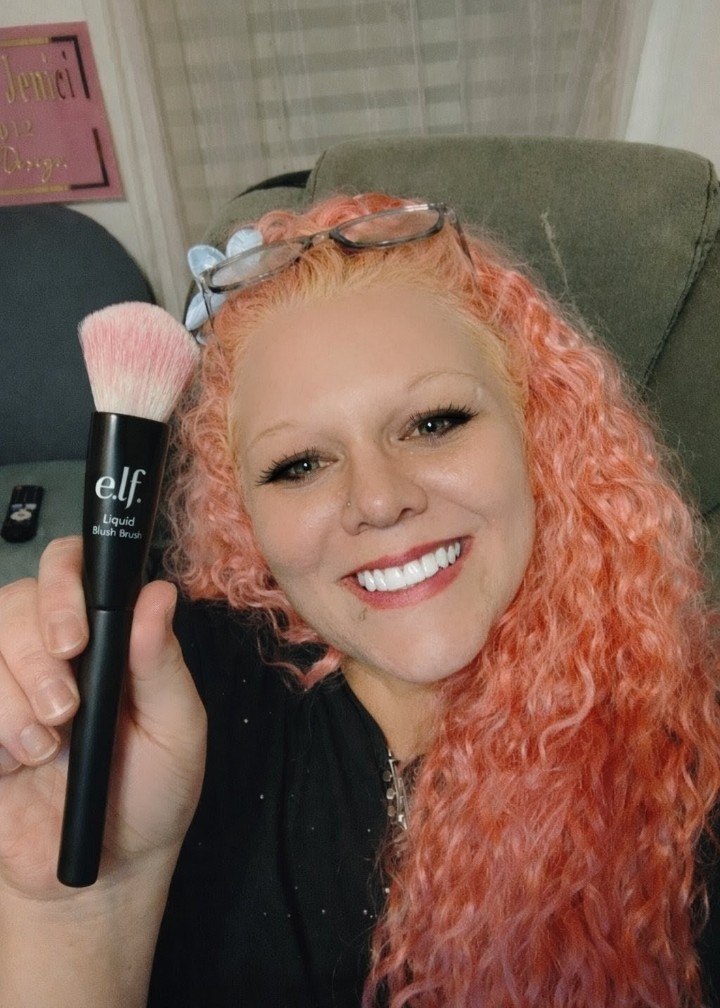 Woman with pink curly hair smiling while holding an e.l.f. Liquid Blush Brush, showcasing the soft pink bristles in a relaxed home environment.
