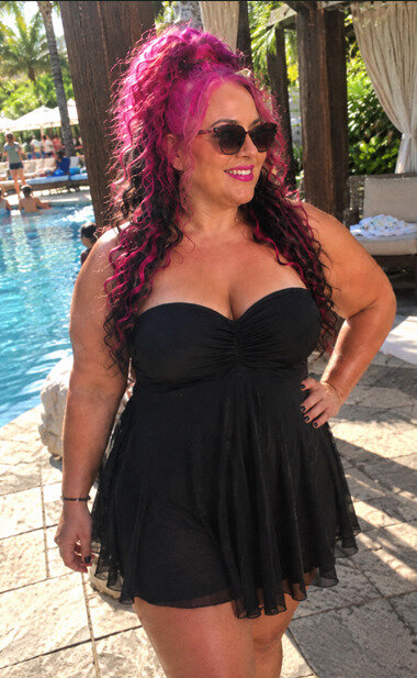Woman with pink and black hair and sunglasses in a black strapless swim dress standing next to a gazebo by a pool.
