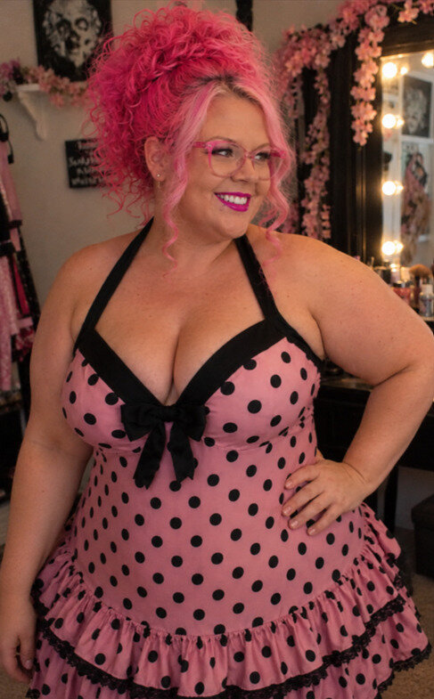 Woman modeling a pink polka dotted swim dress with front bow in front of a vanity mirror.