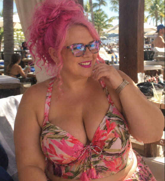 Pink haired woman in a longline swimsuit top in front of a vanity mirror.