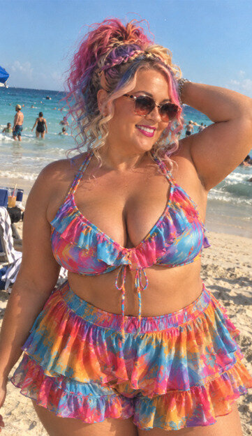 Woman with sunglasses poses in a tie dye shortini with ruffles standing on the beach in front of the ocean.