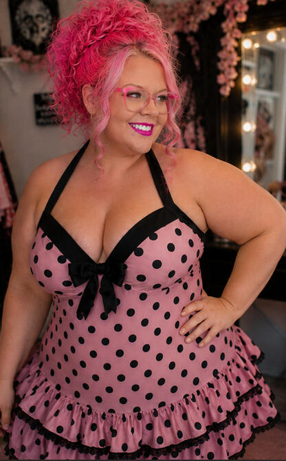 Woman modeling a pink polka dotted swim dress with front bow in front of a vanity mirror.