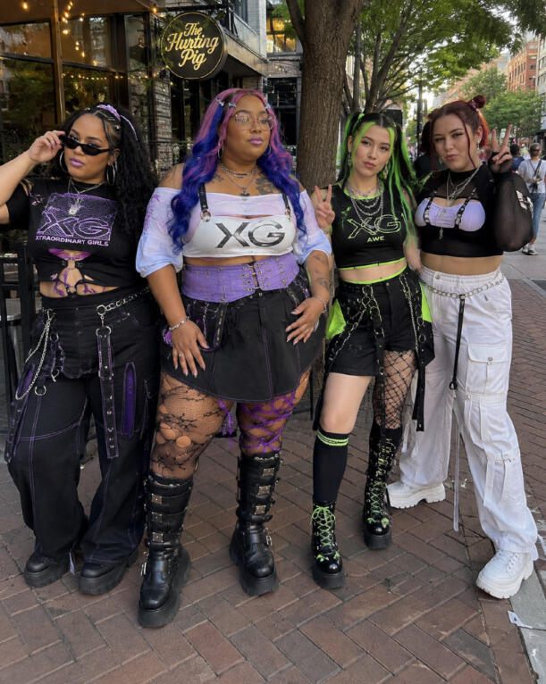 Four women in XG concert outfit ideas posing outdoors on sidewalk.